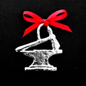 May include: A silver metal ornament shaped like an anvil with a hammer on top. The ornament is decorated with a red bow.