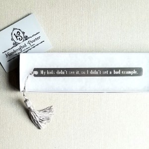 May include: A pewter bookmark with the text "My kids didn't see it, so I didn't set a bad example." The bookmark has a tassel and is packaged in a white box. A card with the text "Handcrafted Pewter" is also visible.