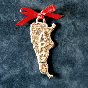 May include: A gold-colored metal ornament in the shape of Argentina, with the letters "ARG" in the center. A red ribbon is tied at the top. The ornament is likely a souvenir or decorative item.