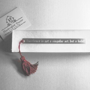 May include: Silver bookmark with a red tassel and the quote "Excellence is not a singular act, but a habit."