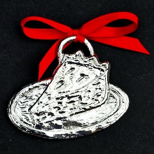May include: A silver-colored pie slice ornament on a plate, with a red ribbon tied at the top. The ornament has a textured surface and is set against a black background. The pie slice has a bite taken out of it.