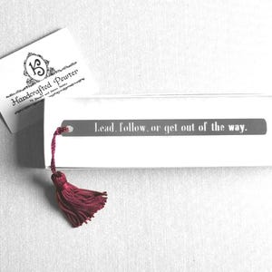 May include: A white bookmark with the text "Lead, follow, or get out of the way." and a burgundy tassel. The bookmark is in a white box with a card that reads "Handcrafted Pewter".