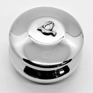 May include: A polished silver-colored round container with a lid. The lid features a raised emblem of a dove in flight. The container has a reflective surface, and the dove emblem is in the center of the lid.