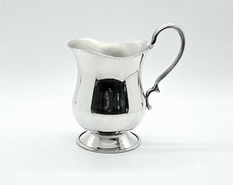 May include: A polished silver-toned creamer pitcher featuring a curved handle, a wide pouring spout, and a rounded body that tapers toward a circular pedestal base. The metallic surface is reflective, showing smooth contours and a classic design.