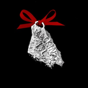 May include: Silver ornament shaped like Mackinac Island with a red ribbon attached. The ornament says "Mackinac Isl."