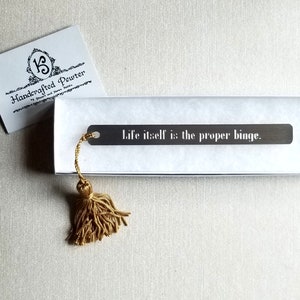 May include: A handcrafted pewter bookmark with the quote "Life itself is the proper binge." The dark gray bookmark has a gold tassel and is presented in a white box. A business card with the logo "Handcrafted Pewter" is also visible.