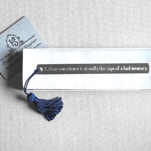 May include: A silver bookmark with a blue tassel and the quote "A clear conscience is usually the sign of a bad memory." The bookmark is on a white surface.