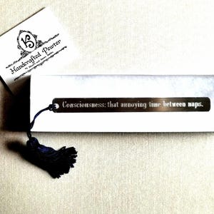 May include: A silver bookmark with a black tassel. The bookmark has the text "Consciousness: that annoying time between naps." engraved on it.