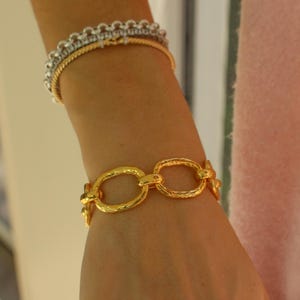 May include: A gold-toned bracelet with textured oval links is the focal point, complemented by a delicate gold and silver bracelet above. Several rings, including a stackable design, adorn the hand. The jewelry is set against a soft pink background.