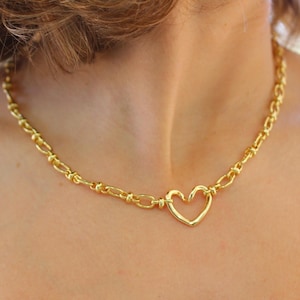 May include: Gold-toned necklace featuring a heart-shaped pendant. The necklace has a chain-link design with a heart pendant at the center. The jewelry is displayed on a person's neck.