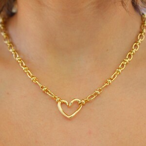 May include: A gold-colored necklace featuring a heart-shaped pendant. The necklace has a chain-link design with a heart pendant in the center. The necklace is a bright gold color and is worn around the neck.