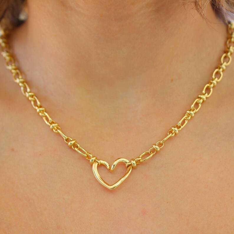 May include: A gold-colored necklace featuring a heart-shaped pendant. The necklace has a chain-link design with a heart pendant in the center. The necklace is a bright gold color and is worn around the neck.