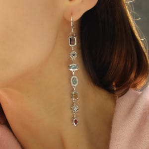 May include: A long, dangling earring featuring a sequence of colourful gemstones in diverse shapes. The gemstones include rectangular, oval, and heart-shaped stones in shades of purple, blue, green, and orange. The earring is made of silver-coloured metal.