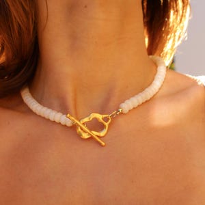 May include: A white beaded necklace with a gold-colored clasp. The clasp has an abstract shape with a bar closure. The necklace is worn around the neck.