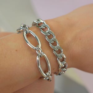 May include: Two silver bracelets are displayed on a wrist. One bracelet features large oval links, while the other has a curb chain design. The bracelets are made of a shiny, metallic material, reflecting light.