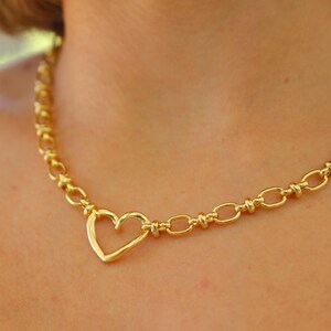 May include: A gold-colored necklace featuring a heart-shaped pendant. The necklace has a chain-link design with a heart pendant at the center. The necklace is worn around the neck.