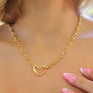 May include: Gold-toned necklace featuring a heart-shaped pendant. The necklace has a chain-link design and is worn around the neck. The heart pendant is the focal point, adding a touch of romance to the jewelry piece. The necklace is a stylish accessory.