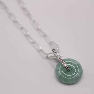 May include: A silver chain necklace with a jade pendant. The pendant is a round disc with a swirl design and is accented with a silver bar set with clear cubic zirconia stones.