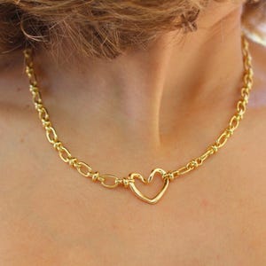 May include: Gold-colored necklace featuring a heart-shaped pendant. The necklace has a chain-link design with a heart-shaped pendant in the center. The necklace is made of a shiny, gold-colored metal. The necklace is worn around the neck.
