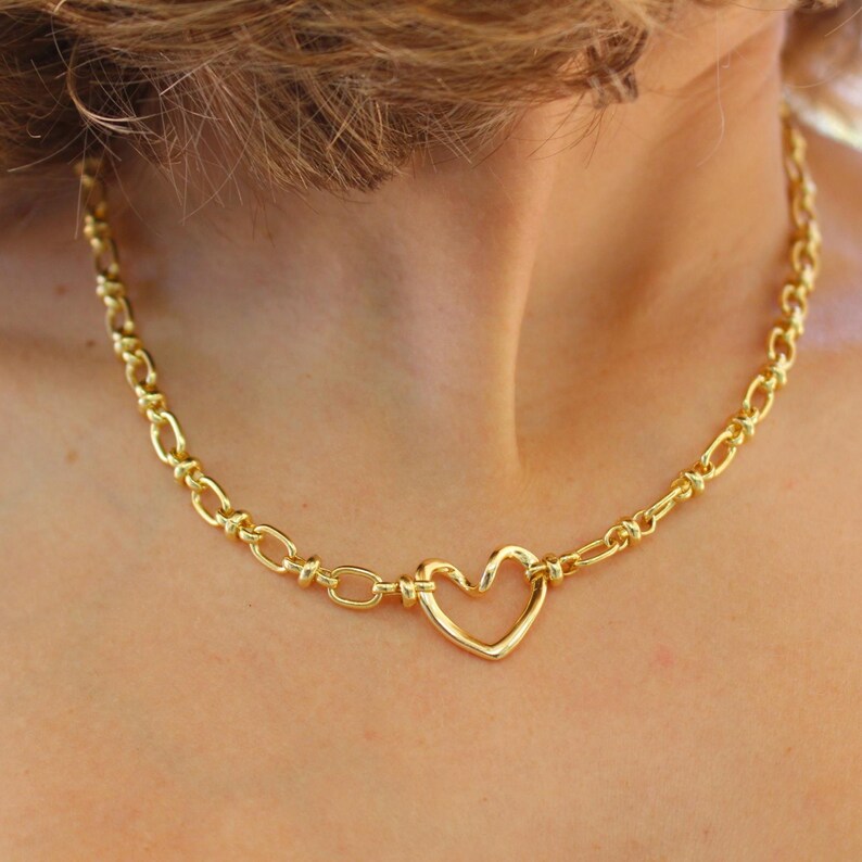 May include: Gold-colored necklace featuring a heart-shaped pendant. The necklace has a chain-link design with a heart-shaped pendant in the center. The necklace is made of a shiny, gold-colored metal. The necklace is worn around the neck.