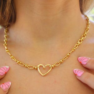 May include: A gold-colored necklace featuring a heart-shaped pendant. The necklace has a chain-link design with a heart-shaped centerpiece. The jewelry is displayed on a person's neck.