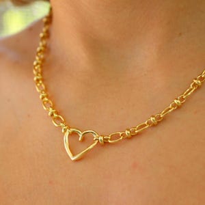 May include: A gold-colored necklace featuring a heart-shaped pendant. The necklace has a chain-link design with a heart-shaped clasp. The jewelry is worn around the neck and is a close-up shot.