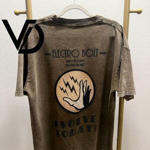 Könnte beinhalten: Ein braunes T-Shirt im Acid-Wash-Look mit einem Grafikdruck auf der Rückseite. Der Text lautet ELECTRO BOLT, DON'T BE A DOLT USE ELECTRO BOLT und EVOLVE TODAY! Ein kreisförmiges Emblem zeigt eine Hand, die nach Blitzen greift, in Schwarz und Beige.