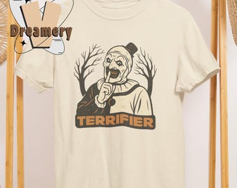 May include: Cream-colored t-shirt featuring a graphic of a scary clown with the word "TERRIFIER" in brown block letters. The clown is depicted with a finger to its lips, with bare tree branches in the background.