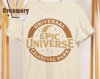 May include: Cream-colored t-shirt with a brown circular design featuring the words "UNIVERSAL EPIC UNIVERSE CELESTIAL PARK" in a vintage style. The design includes a crescent moon and stars. The shirt is hanging on a wooden hanger.