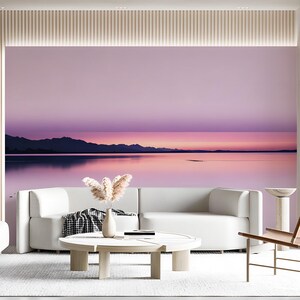 May include: A wall mural featuring a serene landscape of a calm lake reflecting a pink and purple sunset with distant mountain silhouettes. The scene is displayed behind a white curved sofa, a round wooden coffee table, and a small vase of dried grass.