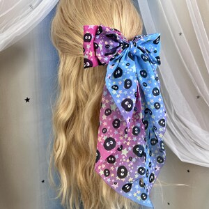 May include: A large hair bow with a colorful design. The bow features a gradient of pink, purple, and blue, adorned with a pattern of black, round characters and white stars. The bow is attached to a blonde wig.