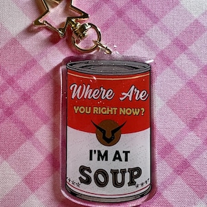Może przedstawiać: Przezroczysty akrylowy brelok z motywem puszki z zupą. Tekst na puszce brzmi "Where Are You Right Now? I'm at Soup." Brelok ma złotą zapięcie w kształcie gwiazdy i złoty kółko na klucze.