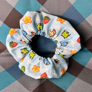 May include: A light blue fabric hair scrunchie featuring a repeating pixel art pattern of small red strawberries, yellow stars, green creatures, and white clouds. The scrunchie has a gathered, ruffled texture and a circular shape.