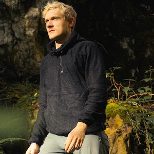 Peut inclure: Un homme portant un sweat-shirt à capuche noir avec une fermeture éclair et un petit drapeau sur la poitrine. Il se tient dans une forêt avec une végétation verte et des rochers.