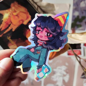 Peut inclure: Autocollant holographique représentant un personnage de dessin animé aux cheveux bleu foncé, vêtu d'une combinaison bleu sarcelle et d'un chapeau de fête. Le personnage tient une bouteille bleue. Finition irisée.