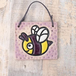 May include: A square ceramic tile with a cartoon bee design. The bee has yellow and purple stripes, white wings, and a black outline. The tile is light purple with small, raised dots and a dark gray leather cord for hanging.