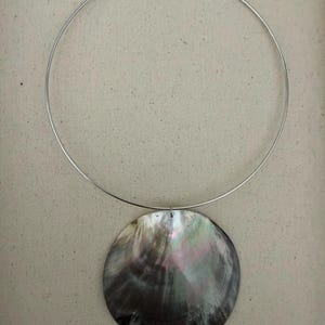 May include: A silver-toned metal choker necklace featuring a large, circular, iridescent shell pendant. The pendant displays dark grey, black, and subtle purple tones with a natural pearlescent sheen. The necklace has a simple hook closure at the top.
