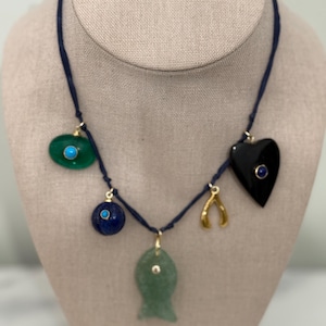May include: A necklace with a dark blue cord and multiple charms. Charms include a green oval with a turquoise stone, a blue sphere, a green fish, a gold wishbone, and a black heart with a blue stone. The necklace is displayed on a mannequin.