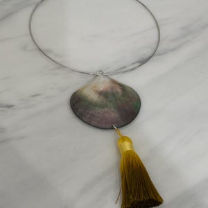 May include: A silver-toned metal choker necklace featuring a large, iridescent seashell pendant in shades of purple, green, and white. A long, mustard-yellow fabric tassel is attached to the bottom of the shell, hanging vertically from the center.