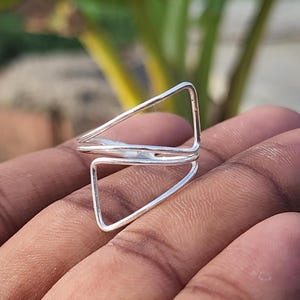 Puede incluir: Un anillo de plata con un diseño geométrico que presenta dos triángulos abiertos. El anillo se sostiene en una mano, mostrando su estilo minimalista y moderno. El diseño del anillo es simple pero llamativo.