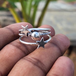 May include: A set of silver rings with star and ball designs. The rings are stacked together, with one ring featuring a star, another with a ball, and others with textured bands. The rings are displayed against a blurred background of green foliage.