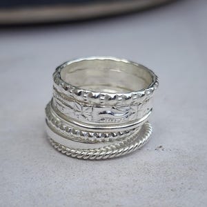 Puede incluir: Una pila de anillos de plata con varias texturas. Los anillos incluyen un diseño de cuerda trenzada, patrones florales y bandas lisas. Los anillos se muestran sobre una superficie de color claro, con un fondo borroso.