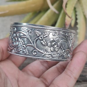May include: A silver cuff bracelet with an intricate floral design. The bracelet is made of metal and has a polished finish.
