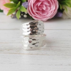 May include: Two silver rings with a hammered texture are stacked on a white wooden surface. The rings are in focus, with a blurred background of pink and white flowers. The rings are a simple, yet elegant design.