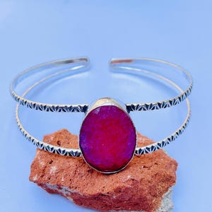 Può includere: Un bracciale rigido in argento con design a doppio cinturino, dettagli incisi e una grande pietra preziosa color magenta sfaccettata di forma ovale incastonata al centro. Il cinturino in metallo ha una finitura lucida e un'apertura posteriore.