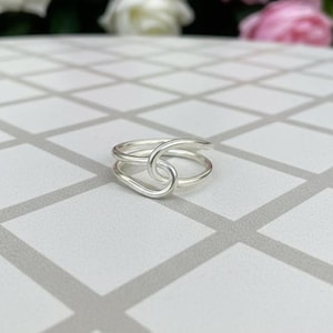 May include: A silver ring with a unique, intertwined design. The ring features a looped upper band that connects to a lower band, creating a continuous, flowing shape. The ring is set against a patterned surface, with soft-focus flowers in the background.