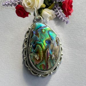Könnte beinhalten: Abalone-Muschel-Anhänger mit silberfarbenem Metallrahmen. Die schillernde Muschel zeigt Farbtöne von Grün, Blau und Lila. Der Anhänger hat einen dekorativen Rand und eine Schlaufe für eine Kette. Rote und weiße Rosen und Lavendelzweige sind im Hintergrund.