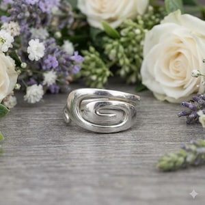 May include: A silver ring featuring a swirling spiral design. The ring is displayed against a backdrop of white roses and purple flowers. The ring's design is the focal point, with a natural aesthetic.