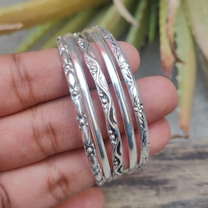 May include: A set of five silver bangles with intricate floral and geometric patterns. The bangles are stacked together, showcasing the detailed designs and the reflective quality of the metal. The jewelry is displayed against a natural background.