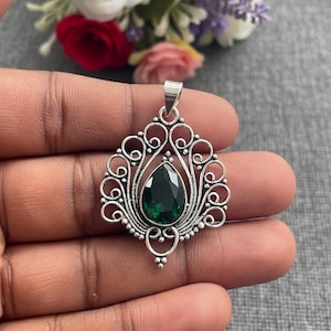 May include: A silver pendant with an ornate, openwork design. The pendant features a pear-shaped, deep green gemstone at its center. The pendant is held in a hand, with a blurred floral arrangement in the background.